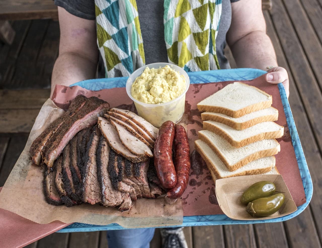 A tray of smoked brisket, sausage, bread, pickles, and potato salad on a rustic porch